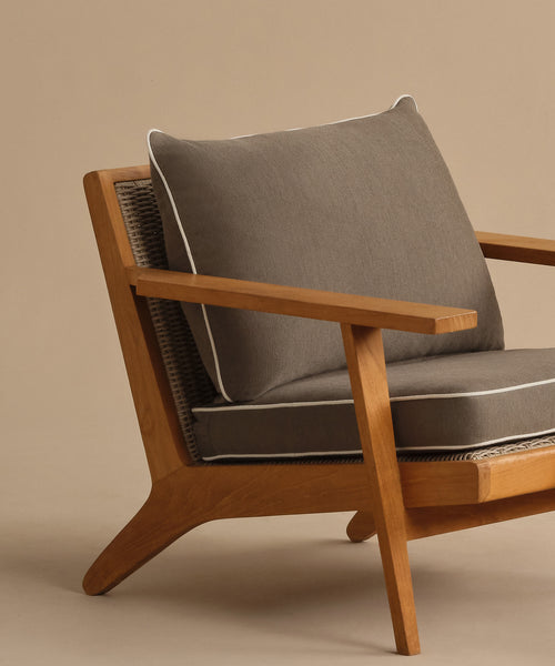 The Outdoor Vista Chair features modern teak wood, angled legs, a woven backrest, and thick gray cushions with white piping, shown against a plain beige background.