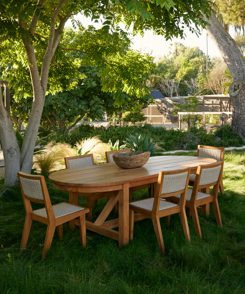 Six Outdoor Vista Dining Chairs and a wooden dining table are arranged on a grassy lawn beneath leafy trees, surrounded by lush greenery and dappled sunlight. A large bowl serves as the tables centerpiece.