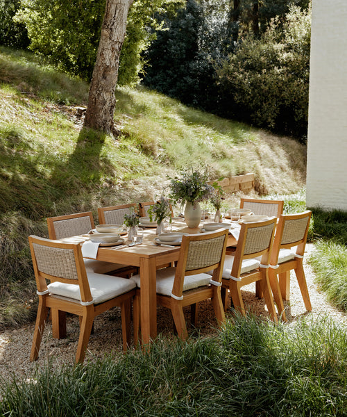 The Outdoor Vista Dining Table with eight cushioned chairs sits on gravel among tall grass and trees, adorned with white napkins, plates, and vases of fresh flowers.