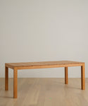 The Outdoor Vista Dining Table, featuring a rectangular solid teak top and four straight legs, sits on a light wood floor against a plain, light-colored wall.