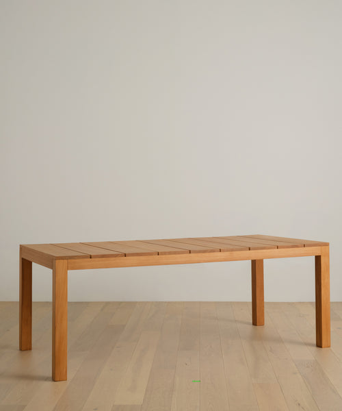 A simple rectangular wooden table with four legs sits on a light wood floor against an off-white wall, protected by the Outdoor Vista Dining Table Cover.