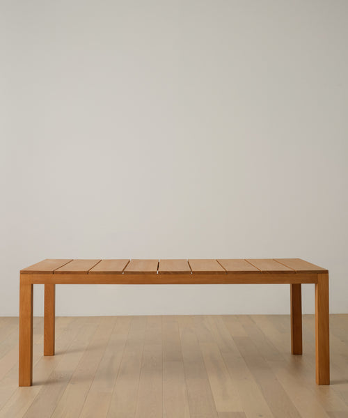 The Outdoor Vista Dining Table, crafted from solid teak with four legs and a slatted top, stands on a light wood floor against a plain white wall.
