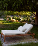 The Outdoor Vista Lounge Chair, featuring a cushion and pillow, is placed on a wooden deck in a shaded garden filled with greenery and sunlit plants beneath a large tree.