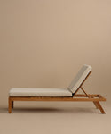 The Outdoor Vista Lounge Chair features a minimalist teak frame and light beige cushions, set against a neutral beige background. Its angled backrest offers comfortable reclining, making it an inviting and durable outdoor furniture piece.