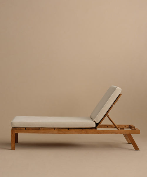 The Outdoor Vista Lounge Chair, featuring a minimalist teak frame and light beige cushions, rests against a plain beige background. Its gently reclined backrest and sturdy beam provide both style and durability for outdoor lounging.