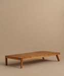 The Outdoor Vista Lounge Chair features a low, rectangular teak design with a slatted top and angled legs, displayed against a plain beige background.