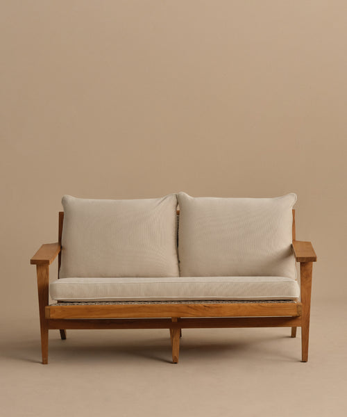The Outdoor Vista Sofa features a wooden frame, light beige cushions, and two large back pillows, displayed against a plain beige background.