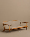 The Outdoor Vista Sofa features a minimalist design with angled legs, wide armrests, and a woven beige back and seat, shown against a plain light brown background.