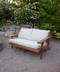 The Outdoor Vista Sofa, featuring Tempotest fabric cushions, sits on sandy ground amid rocks, green bushes, and blooming white flowers.