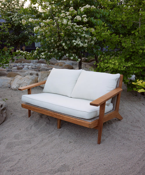 The Outdoor Vista Sofa, featuring Tempotest fabric cushions, sits on sandy ground amid rocks, green bushes, and blooming white flowers.