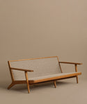 The Outdoor Vista Sofa features a minimalist teak frame with angled legs and arms, plus a woven beige seat and backrest. Shown on a plain beige background, its even comfier and more durable with Tempotest fabric cushions.