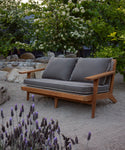 The Outdoor Vista Sofa with gray cushions is placed on sandy ground in a garden, surrounded by blooming flowers, leafy green trees, and large rocks.