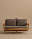 The Outdoor Vista Sofa features a teak wooden frame, angled armrests, grey seat and back cushions, and a beige backdrop. Its minimalist, modern design makes it ideal for adding style to any patio space.