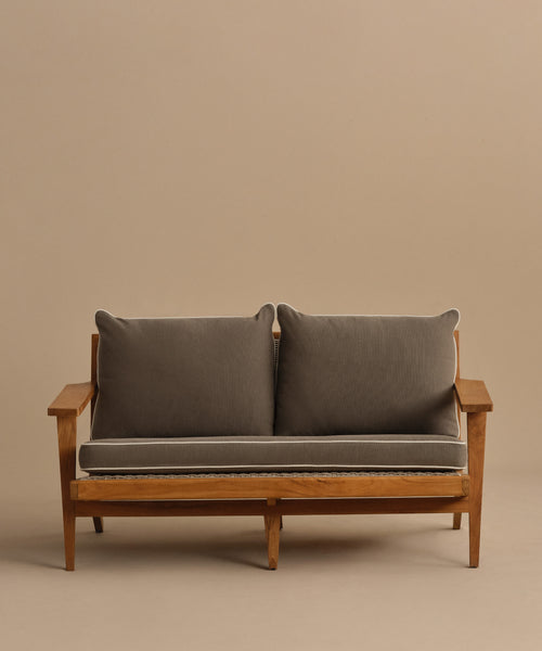 The Outdoor Vista Sofa features a teak wooden frame, angled armrests, grey seat and back cushions, and a beige backdrop. Its minimalist, modern design makes it ideal for adding style to any patio space.