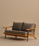 The Outdoor Vista Sofa features modern teak with angled legs, wide armrests, and two gray cushions for seat and back—ideal ECOLENE furniture for any contemporary space against a plain beige background.