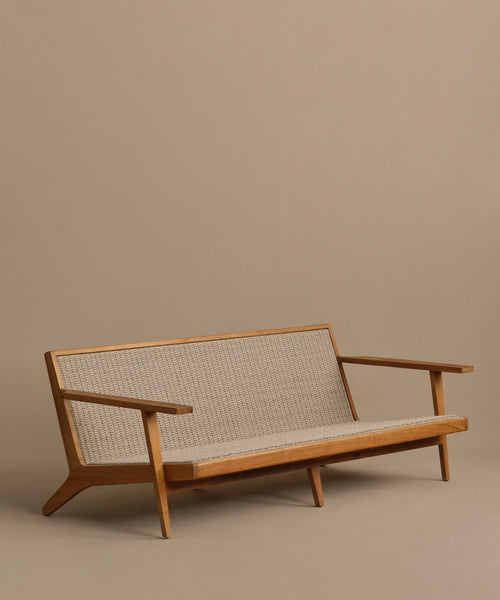 The Outdoor Vista Sofa is a modern, minimalist teak piece with angled legs and armrests, featuring a woven beige ECOLENE seat and backrest, set against a plain beige background.