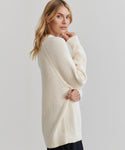A woman with blonde hair wears the Oversized Cotton Fisherman sweater and dark pants, standing sideways and looking over her shoulder against a plain light background.