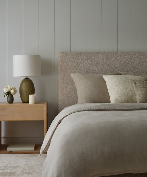 The Pacific Bed, featuring natural wool boucle and an upholstered headboard with Belgian linen bedding, is placed beside a wooden nightstand holding a lamp, white flowers in a vase, and a candle against a light gray paneled wall.