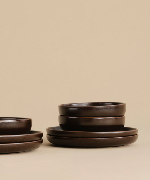The Pacific Breakfast Bowl, a dark chocolate-colored stoneware piece, sits among matching bowls and plates on a light beige surface with a coordinating background.