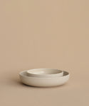 Two minimalist, round Pacific Breakfast Bowls in off-white—one nested inside the other—rest elegantly on a beige surface, set against a beige background. Modern dinnerware for any setting.
