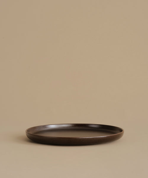 A dark brown ceramic dinner plate with a matte finish, photographed against a solid beige background.
