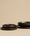 The Pacific Dinner Plate, crafted from dark brown ceramic stoneware, is showcased with matching bowls against a beige background. The minimalist display emphasizes the smooth, matte texture of this elegant dinnerware set.