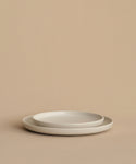 Two simple, round Pacific Dinner Plates in white stoneware are stacked against a beige background. Their minimalist design and soft lighting create a clean, modern look for any setting. Made in Portugal.