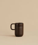 The Pacific Mug, made of dark chocolate-colored stoneware with a handle, rests on a beige surface against a plain beige background.