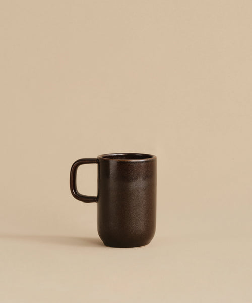 The Pacific Mug, made of dark chocolate-colored stoneware with a handle, rests on a beige surface against a plain beige background.