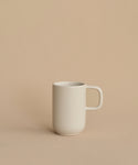 The Pacific Mug, a plain white stoneware piece from Pacific Dinnerware and made in Portugal, sits slightly left of center on a beige surface with a matching background. Simple and unadorned, it offers understated elegance.