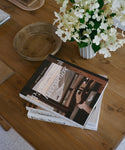 Stack of books on a wooden table with a vase of white flowers and a wooden bowl.