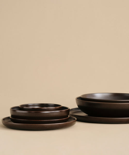 A stack of Pacific Salad Plates in dark brown, arranged on a beige surface with a beige background in two neat piles.