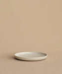 A minimalist, round, white Pacific Salad Plate made of stoneware in Portugal rests empty on a beige surface with a beige background.
