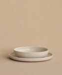 A Pacific Soup Bowl with gently raised sides is stacked on a plain flat plate atop a beige surface. Both white ceramic pieces, made in Portugal, feature a refined and elegant design.