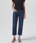 A person in a black short-sleeve top, Parker Jean high-rise vintage denim jeans, and black heeled sandals stands against a white background with their left hand in their pocket.