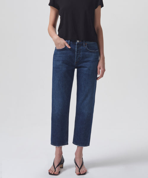A person in a black short-sleeve top, Parker Jean high-rise vintage denim jeans, and black heeled sandals stands against a white background with their left hand in their pocket.