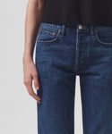 A person wears high-waisted, dark blue Parker Jean by AGOLDE and a black top. The image, cropped from shoulders to upper thighs, shows their relaxed hand at the side against a plain, light background.