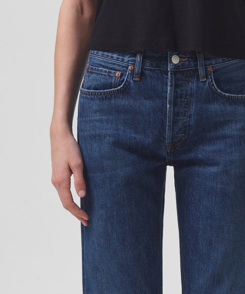 A person wears high-waisted, dark blue Parker Jean by AGOLDE and a black top. The image, cropped from shoulders to upper thighs, shows their relaxed hand at the side against a plain, light background.