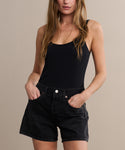 A woman wearing a black sleeveless top and Parker Long Short denim shorts stands with her hands in her pockets against a neutral background.