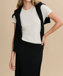 A woman in a white short-sleeve t-shirt, black sweater over her shoulders, and the Penelope Skirt stands against a beige background with one hand on her hip.