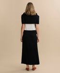 A woman stands with her back to the camera, wearing a white top, black sweater over her shoulders, long black Penelope Skirt, and brown flats against a plain beige background.