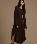 A woman with long dark hair poses against a beige background, wearing a fitted, dark brown ribbed top and the Penelope Skirt—a pleated midi with an elegant asymmetrical hem.