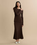A woman with long dark hair stands against a beige background, wearing the Penelope Skirt—a long, ribbed brown knit skirt—with a matching brown ribbed top and white slip-on shoes, her expression neutral.