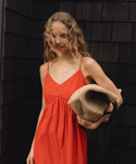 Woman in an red dress holding a beige woven raffia handbag against a dark wooden wall.