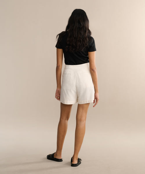 A person with long wavy hair, in a black t-shirt, cream Pleated Short, and black sandals, stands facing away against a neutral beige background.