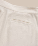 Close-up of Pleated Short in light beige textured fabric, high-waisted with a single welt back pocket and visible seam details, shown against a neutral background.