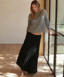 A woman stands barefoot in a bright room with wood floors and glass doors, wearing a gray ribbed knit sweater and a long black pleated skirt.