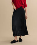 A person stands against a beige background wearing a Pleated Skirt, a red sweater, and black slip-on shoes. The person's face is not visible.