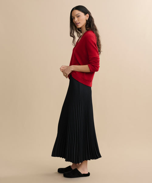 A woman stands sideways against a beige background, wearing a black Pleated Skirt and a red long-sleeve sweater. Her long wavy hair flows freely, she wears black slippers, and she carries a calm expression.