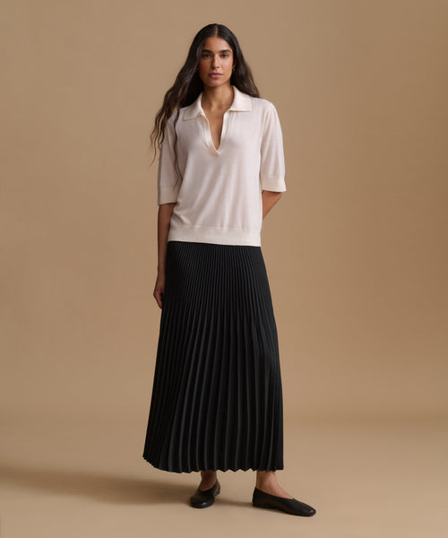 A woman stands against a warm beige studio background wearing a cream short-sleeve collared knit top and a long black pleated skirt, paired with black flats.